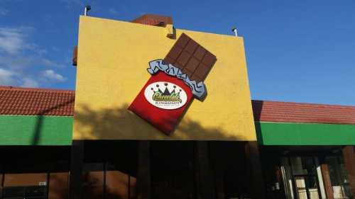 A colorful building exterior featuring a large chocolate bar sign with the name "Chocoholics Kingdom."