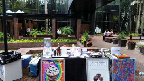 A colorful outdoor setup featuring a chocolate stand with various treats, surrounded by greenery and people in the background.