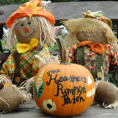 Two scarecrows sit beside a decorated pumpkin labeled "Mrs. Heather's Pumpkin Patch," surrounded by burlap sacks.