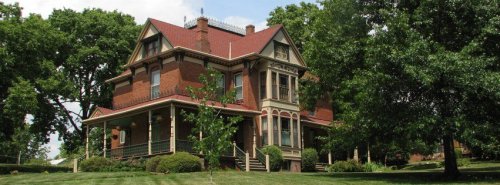 A large, historic brick house with a red roof, surrounded by green trees and a well-maintained lawn.