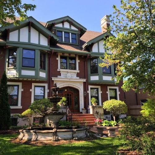 A large, elegant brick house with green trim, a fountain in the front yard, and lush landscaping.