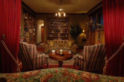 Cozy living room with plaid furniture, a round table, bookshelves, and warm lighting, framed by red curtains.