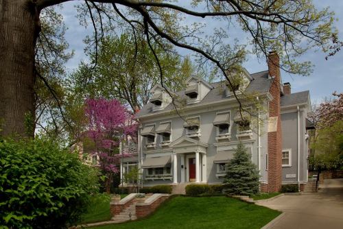 A large, elegant gray house surrounded by greenery and blooming trees on a sunny day.