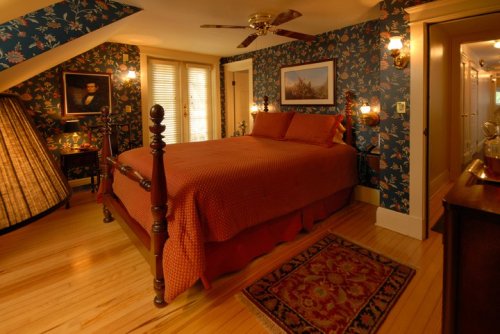 Cozy bedroom with a wooden bed, floral wallpaper, and warm lighting, featuring a rug and artwork on the walls.