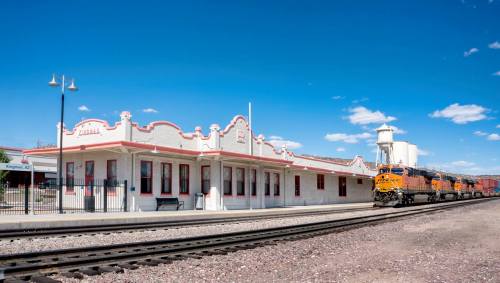 Historic train station with a freight train passing by, set against a clear blue sky.