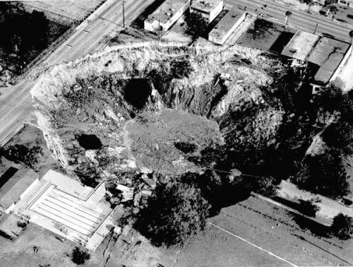 Aerial view of a large, circular crater surrounded by buildings and roads, showing signs of erosion and debris.
