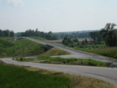 A rural landscape featuring a bridge over a road, surrounded by greenery and distant hills under a clear sky.