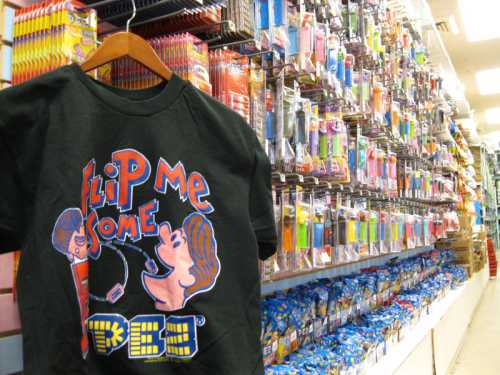 A black T-shirt with a colorful graphic hangs in a store aisle filled with various toys and collectibles.