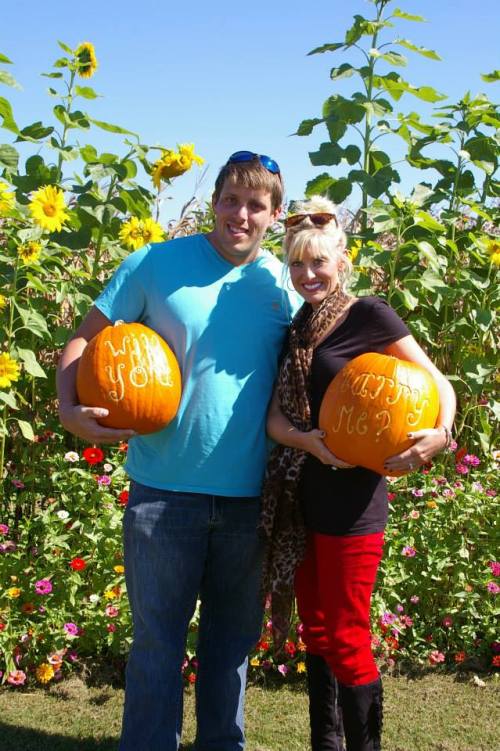 A man and woman hold decorated pumpkins in a flower-filled field, smiling and enjoying a sunny day.