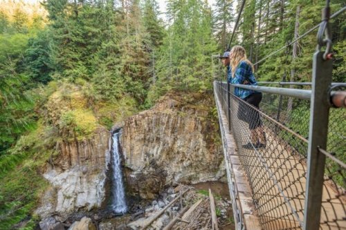 A person stands on a suspension bridge, photographing a waterfall surrounded by lush green trees.