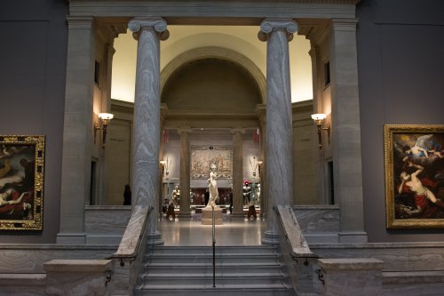 A grand museum entrance with marble columns, stairs, and artwork lining the walls, leading to a central exhibit.
