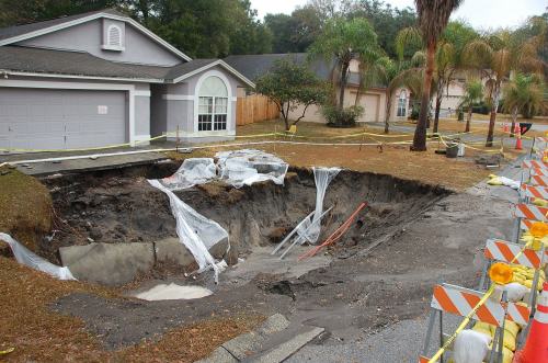 A large sinkhole in a residential area, surrounded by caution tape and construction barriers, near a house.