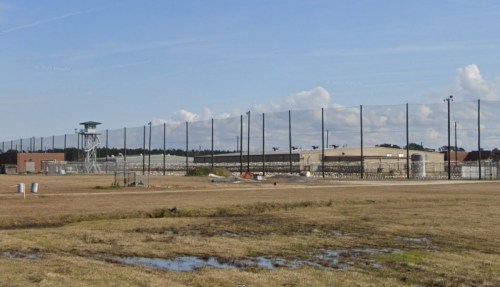 A large prison facility surrounded by tall fences and a watchtower, set against a clear blue sky.