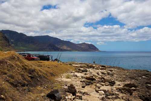 A rocky shoreline with fishing boats and poles, mountains in the background, under a partly cloudy sky.