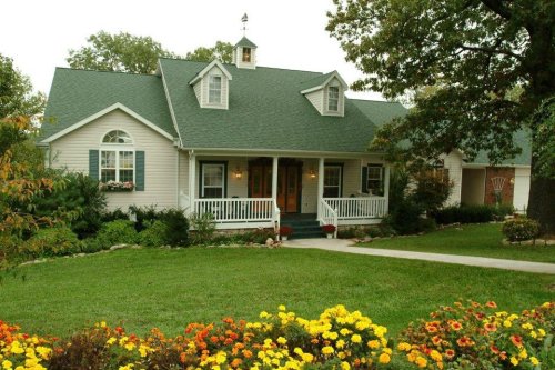A charming house with a green roof, white siding, and a flower garden in front, surrounded by trees.
