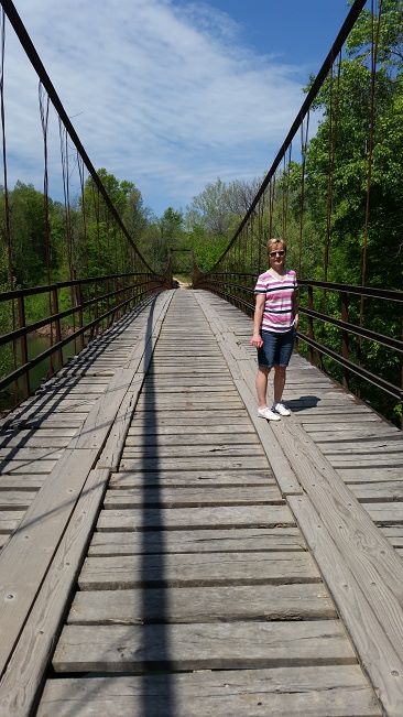 A woman stands on a wooden suspension bridge surrounded by greenery under a clear blue sky.