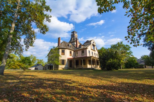A large, historic house with a tower, surrounded by trees and a grassy area under a blue sky with scattered clouds.