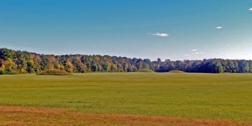 A wide grassy field with two mounds and a backdrop of colorful trees under a clear blue sky.