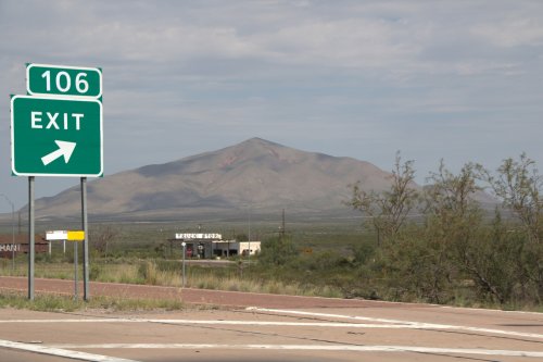 A highway exit sign for "106" with a mountain in the background and a small building nearby.