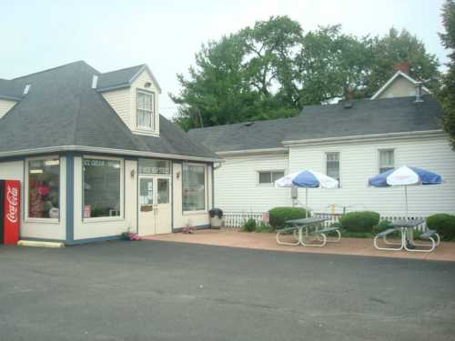 A small ice cream shop with a white exterior, picnic tables, and umbrellas in a parking lot surrounded by greenery.