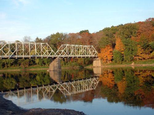A metal bridge spans a calm river, reflecting autumn trees in vibrant colors on the water's surface.