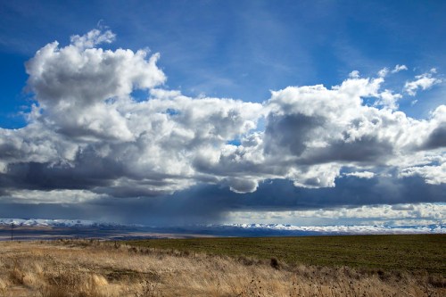 A vast landscape with rolling hills, grassy fields, and a dramatic sky filled with clouds and distant mountains.