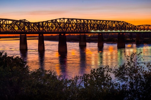 A silhouetted bridge spans a river at sunset, reflecting vibrant colors on the water's surface.