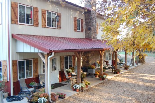 A cozy two-story house with a red metal roof, surrounded by autumn foliage and decorated with potted flowers.
