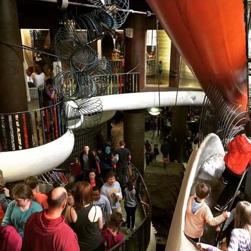 A busy indoor space with a large slide and spiral structures, filled with people enjoying the playful environment.