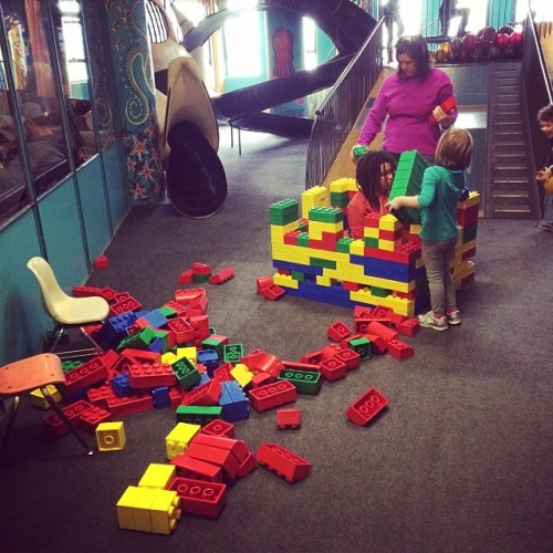 Children playing with colorful building blocks in a play area, with a slide and climbing structure in the background.