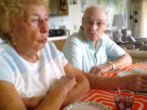 An elderly couple sitting at a table, engaged in conversation, with plates and drinks in front of them.