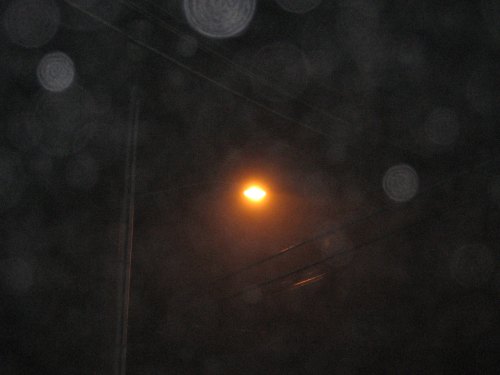 A dimly lit streetlamp shines through a foggy, blurred background with raindrops or mist in the foreground.