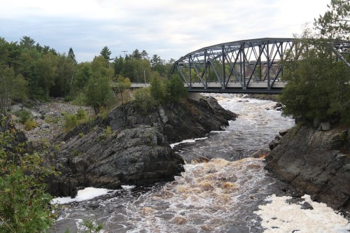 A metal bridge spans a rushing river surrounded by rocky banks and lush greenery under a cloudy sky.