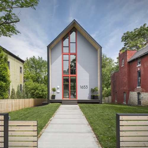 Modern house with a tall, triangular roof, large windows, and a landscaped front yard, flanked by wooden fences.