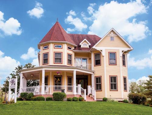 A charming yellow Victorian house with a turret, wraparound porch, and lush green lawn under a blue sky with clouds.