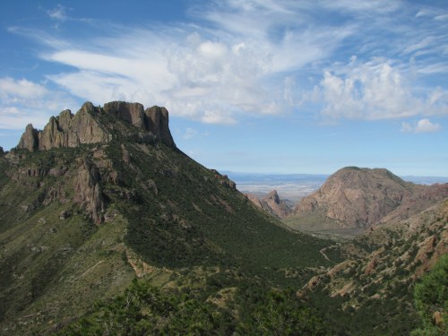 A scenic view of rugged mountains under a blue sky with scattered clouds, showcasing lush greenery and rocky terrain.