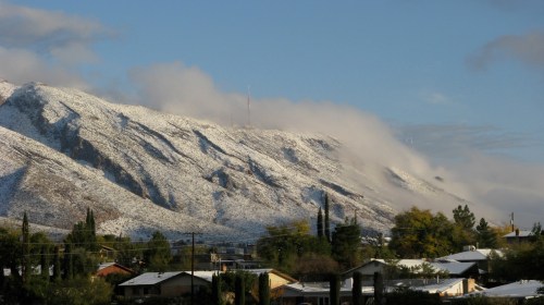 Snow-capped mountains under a cloudy sky, with houses and trees in the foreground.