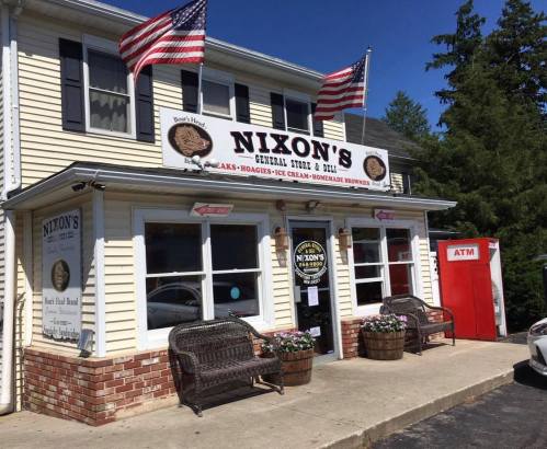 A general store with a sign reading "Nixon's," featuring flags, outdoor seating, and an ATM nearby.