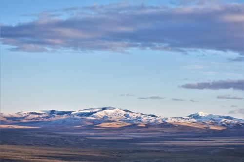 Snow-capped mountains under a blue sky with scattered clouds, overlooking a vast, open landscape.