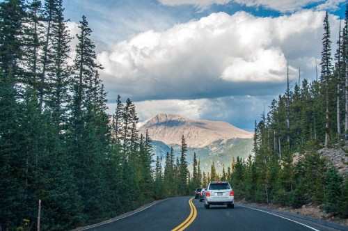 A winding road through a forested area, with mountains and dramatic clouds in the background.