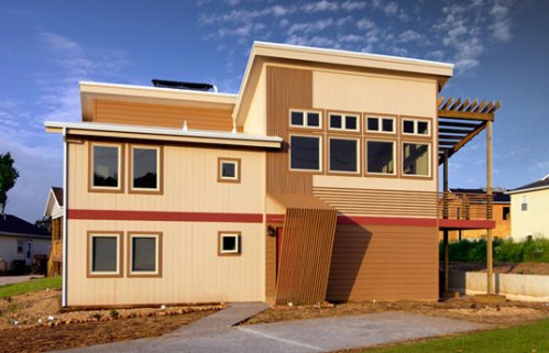 Modern two-story house with a mix of wood and siding, large windows, and a unique angled design. Clear blue sky in the background.