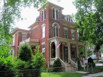 Victorian-style red brick house with a porch, surrounded by greenery and featuring an American flag.