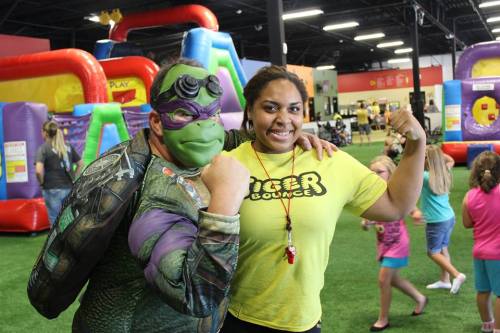 A costumed character and a woman in a yellow shirt flex their muscles at a fun indoor play area with kids in the background.
