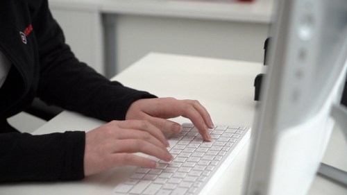 A person typing on a white keyboard at a desk, with a computer monitor in the background.