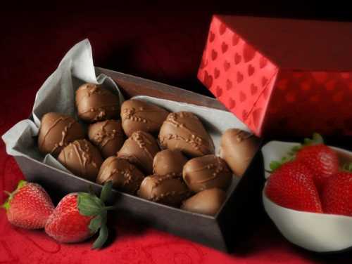 A box of chocolate-covered strawberries next to a bowl of fresh strawberries on a red background.