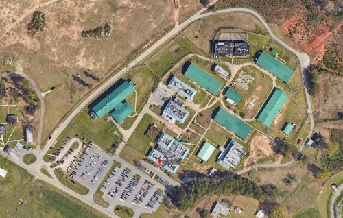 Aerial view of a large facility with multiple buildings and parking areas, surrounded by green fields and trees.
