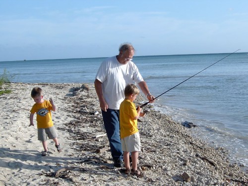 A man fishing on the beach with two young boys, one watching and the other walking along the shore.