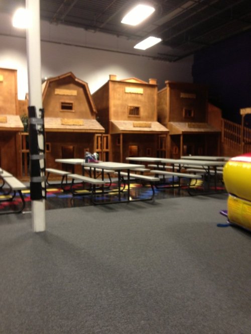 Indoor play area featuring wooden playhouses and tables, with colorful flooring and inflatable structures in the background.