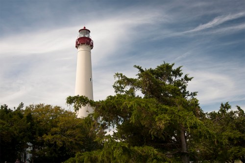 A tall white lighthouse with a red top stands among green trees under a cloudy sky.