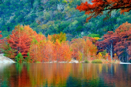 Vibrant autumn foliage reflects in a calm lake, surrounded by lush green hills.